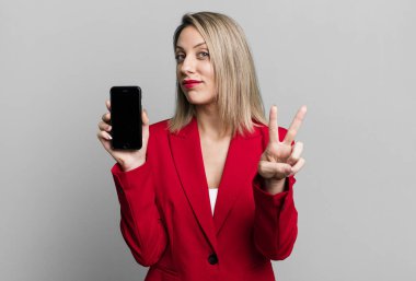 pretty blonde woman smiling and looking happy, gesturing victory or peace. businesswoman showing a phone screen