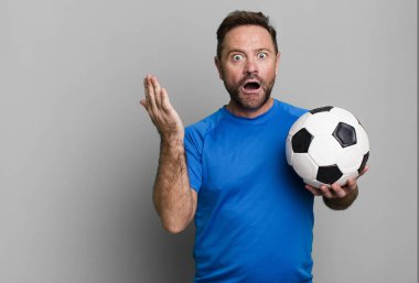 middle age man feeling extremely shocked and surprised. with a soccer ball. fitness concept