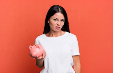 hispanic pretty woman feeling puzzled and confused with a piggy bank