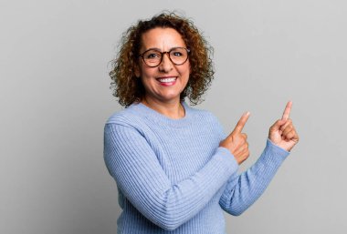 Orta yaşlı İspanyol kadın mutlu bir şekilde gülümsüyor ve iki elini de fotokopi alanında objeler gösteriyor.