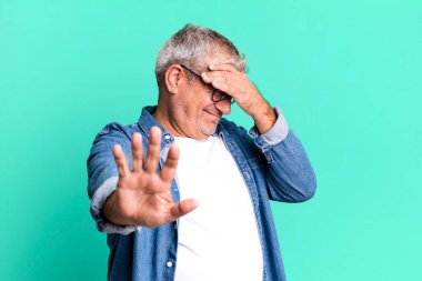 Orta yaşlı bir adam yüzünü elleriyle kapatıyor ve kamerayı durdurmak için diğer elini öne koyuyor, fotoğraf ya da resimleri reddediyor.