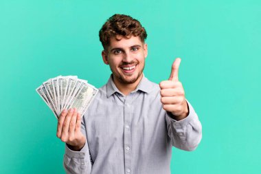 young adult caucasian man feeling proud,smiling positively with thumbs up. dollar bank notes concept