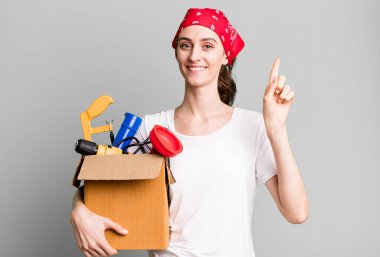 young pretty woman smiling and looking friendly, showing number one. housekeeper and toolbox concept