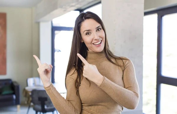 young adult pretty woman smiling happily and pointing to side and upwards with both hands showing object in copy space