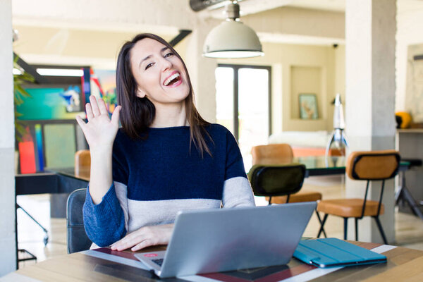 young pretty woman smiling happily, waving hand, welcoming and greeting you. telecommuting concept