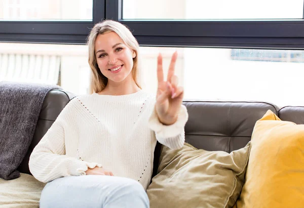 young pretty woman smiling and looking happy, carefree and positive, gesturing victory or peace with one hand sitting on a couch