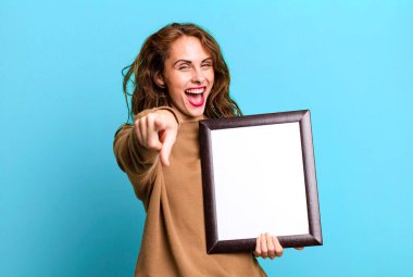 hispanic pretty woman pointing at camera choosing you with an empty blank frame
