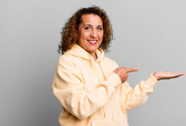 Orta yaşlı İspanyol kadın neşeyle gülümsüyor ve yan taraftaki fotokopi alanını işaret ediyor, bir nesneyi gösteriyor ya da reklamını yapıyor.