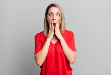 blonde adult woman feeling shocked and scared, looking terrified with open mouth and hands on cheeks