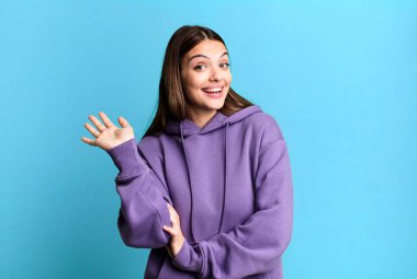 pretty young adult woman smiling happily and cheerfully, waving hand, welcoming and greeting you, or saying goodbye