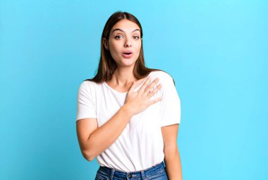pretty young adult woman feeling shocked and surprised, smiling, taking hand to heart, happy to be the one or showing gratitude