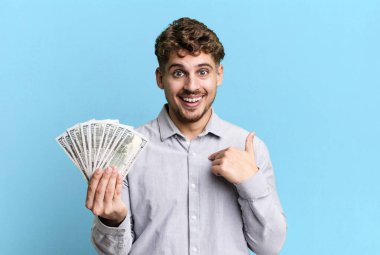 young adult caucasian man feeling happy and pointing to self with an excited. dollar bank notes concept