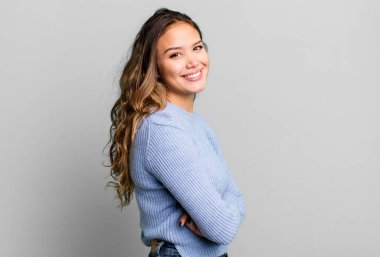 Genç ve güzel bir kadın kameraya çapraz kollarıyla gülümsüyor ve mutlu, kendinden emin, tatmin olmuş bir ifade, yan bakış.