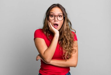Genç ve güzel bir kadın şok ve inançsızlık içinde, elleri yanağında ve kolları çaprazlanmış, sersemlemiş ve şaşırmış hissediyor.