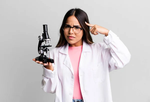 hispanic pretty woman feeling confused and puzzled, showing you are insane. scients student with a microscope