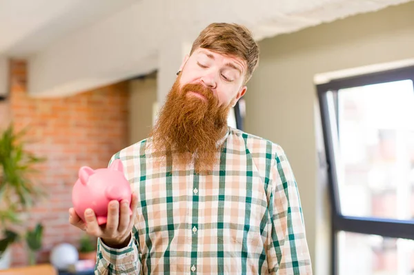 红头发的男人笑着 带着快乐而自信的表情看着小猪银行 — 图库照片