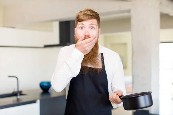 赤い髪の男が手で口を覆いキッチンでショックや驚きの表情 シェフのコンセプト — ストック写真