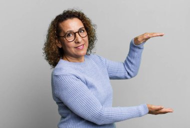 Orta yaşlı İspanyol bir kadın iki eli yanda bir objeyi tutuyor, fotokopi alanında, bir nesneyi gösteriyor, sunuyor ya da reklamını yapıyor.