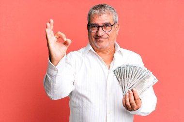 Orta yaşlı bir adam kendini mutlu hissediyor. Onayını iyi bir jestle gösteriyor. Dolar banknotları konsepti