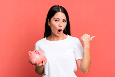 hispanic pretty woman looking astonished in disbelief with a piggy bank