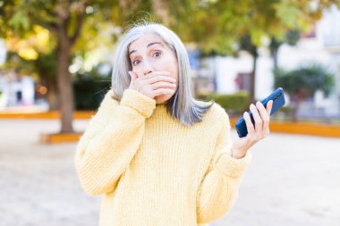 Güzel, son sınıf öğrencisi bir kadın ağzını eliyle kapatıyor ve şok olmuş ya da akıllı telefonla şaşırmış bir ifade takınıyor.