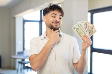 Genç ve yakışıklı bir adam mutlu ve kendinden emin bir ifadeyle gülümsüyor, elleri çenesinde. Dolar banknotları konsepti