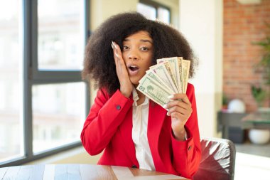 Oldukça afro siyahi bir kadın son derece şok olmuş ve şaşırmış hissediyor. Dolar banknotları konsepti