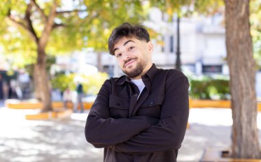 Genç yakışıklı adam omuz silkiyor, kafası karışık ve kararsız, kolları bağlı ve şaşkın görünüyor.