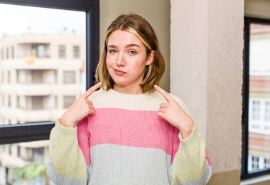 pretty caucasian woman with a bad attitude looking proud and aggressive, pointing upwards or making fun sign with hands. home interior concept