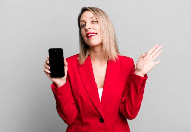 Güzel sarışın kadın mutlu bir şekilde gülümsüyor, el sallıyor, hoş geldin diyor ve seni selamlıyor. İş kadını telefon ekranı gösteriyor