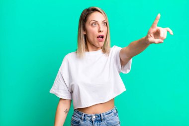 blonde adult woman feeling shocked and surprised, pointing and looking upwards in awe with amazed, open-mouthed look