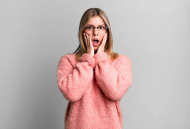 Sarışın, güzel bir kadın şok olmuş ve korkmuş hissediyor, ağzı açık ve elleri yanaklarında korkmuş görünüyor.