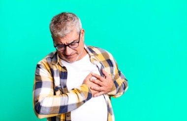 Orta yaşlı, üzgün ve kalbi kırık görünen, iki elini de kalbe yakın tutan, ağlayan ve depresyonda hisseden bir adam.