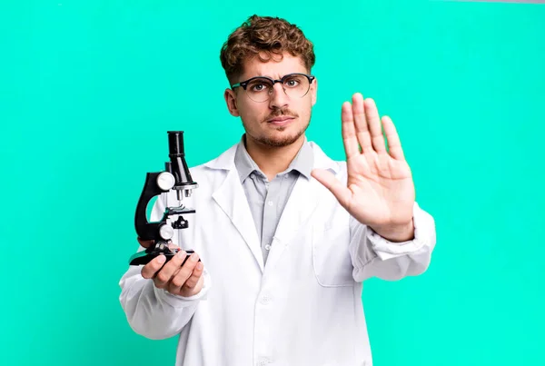 年轻的高加索男子看上去很严肃 表现出开放的手心制造停止动作 带有显微镜概念的科学实验室学生 — 图库照片