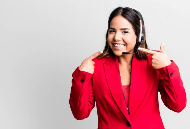 hispanic pretty woman smiling confidently pointing to own broad smile. telemarketing concept