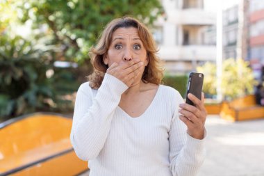 Orta yaşlı, güzel bir kadın ağzını eliyle kapatıyor ve şok olmuş ya da şaşırmış bir ifade takınıyor. Akıllı telefonunu kullanarak.
