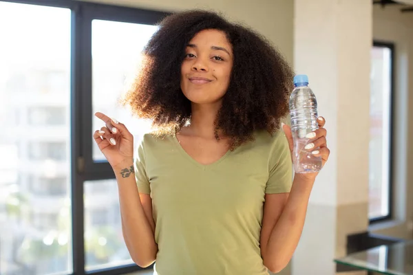 stock image pretty afro black woman smiling cheerfully, feeling happy and pointing to the side. water bottle concept