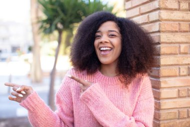Afro-amerikan güzel kadın neşeyle gülümsüyor ve bir objeyi göstererek ya da reklamını yaparak avucundaki kopya alanını işaret ediyor.