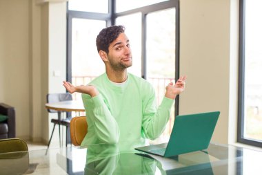 Genç İspanyol adam şaşkın ve şaşkın hissediyor, şüpheci, ağırlıklı ya da farklı seçenekler seçiyor. evde çalışmak