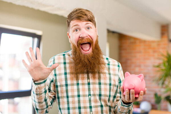 red hair man feeling happy and astonished at something unbelievable with a piggy bank