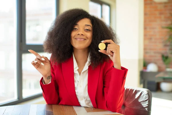 pretty afro black woman smiling cheerfully, feeling happy and pointing to the side. virtual coin concept