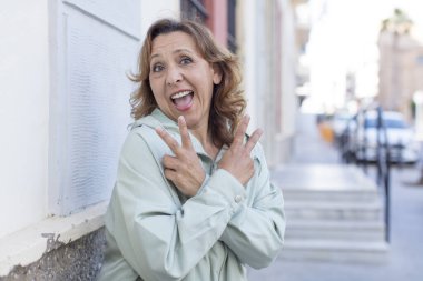 Orta yaşlı kadın gülümsüyor ve mutlu görünüyor, arkadaş canlısı ve memnun, zafer ya da barış işareti yapıyor iki eliyle