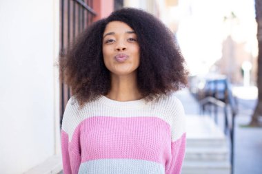 Afro-Amerikan güzel kadın dudaklarını tatlı, eğlenceli, mutlu, sevimli bir ifadeyle birbirine bastırıyor. Öpücük yolluyor.