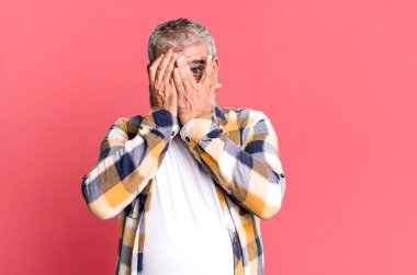Orta yaşlı bir adam elleriyle yüzünü kapatıyor, parmaklarının arasından şaşkın bir ifadeyle yan tarafa bakıyor.