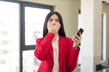 Güzel genç bir kadın ağzını eliyle kapatıyor ve şok olmuş ya da şaşırmış bir ifade takınıyor. akıllı telefon kavramı