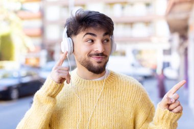 Genç yakışıklı adam neşeyle gülümsüyor, mutlu hissediyor ve bir tarafı işaret ediyor. kulaklıkla müzik dinlemek
