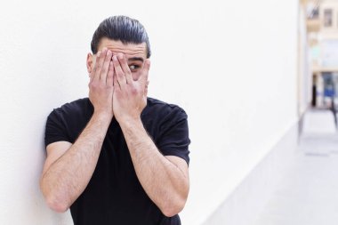 Havalı genç adam elleriyle yüzünü kapatıyor, parmaklarının arasından şaşkın bir ifadeyle yan tarafa bakıyor.