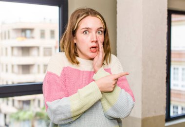 pretty caucasian woman feeling surprised with a shocked expression and pointing to the side. home interior concept