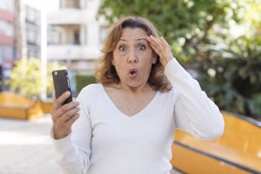 Orta yaşlı, güzel bir kadın mutlu, şaşkın ve şaşırmış görünüyor. Akıllı telefonunu kullanarak.