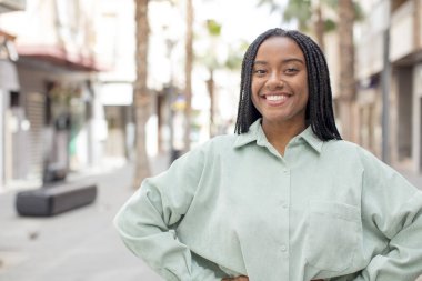 Afro güzel siyah kadın mutlu bir şekilde gülümsüyor. Elleri kalçasında, kendinden emin, gururlu ve arkadaş canlısı bir tavırla.